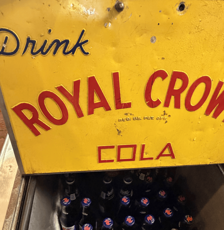 Yellow sign with 'Drink Royal Crown Cola'; bottles below.