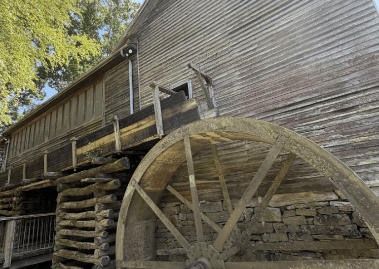 Old wooden watermill with large weathered wheel, under a clear sky.