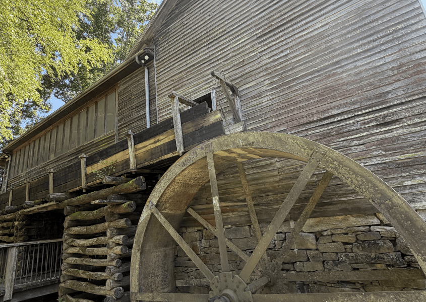 Old wooden watermill with large weathered wheel, under a clear sky.
