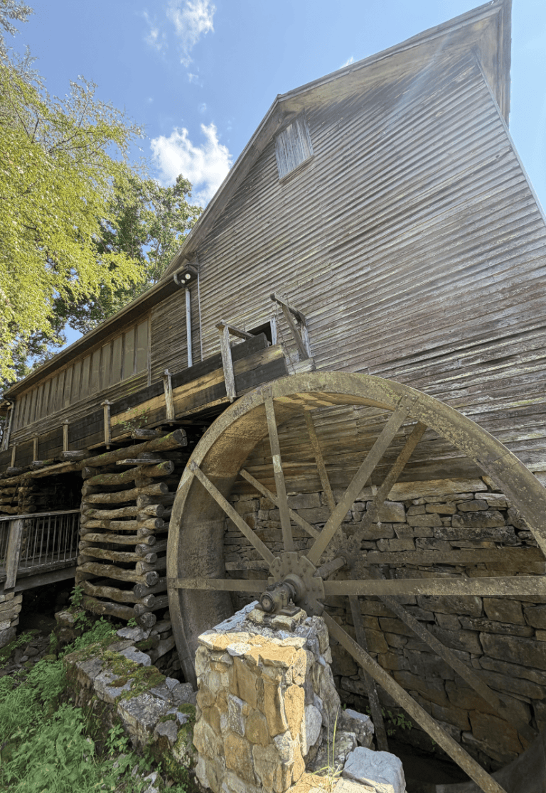 Old wooden watermill with a large wheel and stone foundation under a clear sky.