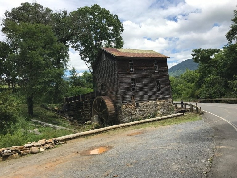 Old watermill by a road surrounded by trees and mountains.