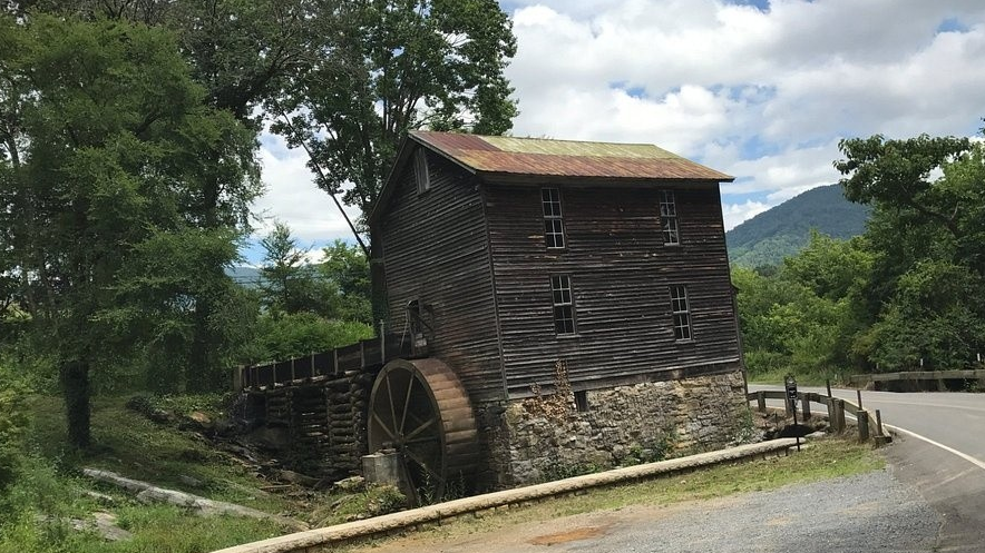 Old wooden mill with waterwheel by a curving road and trees.